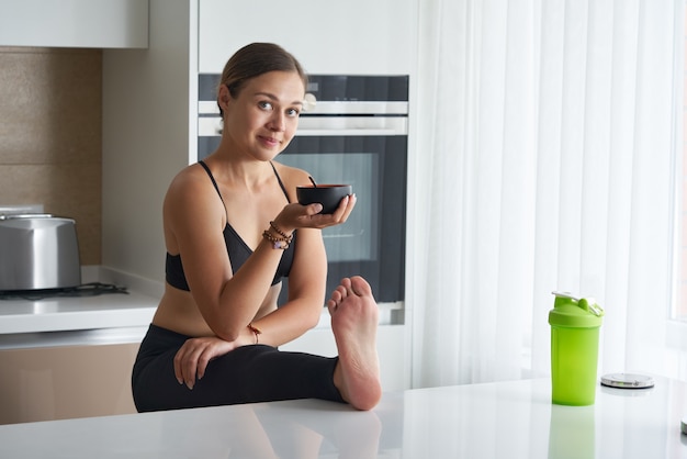 Young woman smiling and eating while stretching in kitchen with leg on table