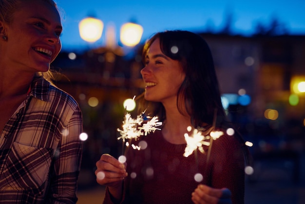 Young woman smiling at her friend and celebrating playing with fireworks Two friends on holiday together celebrating with fireworks at nightHappy woman on holiday with friend playing with fireworks