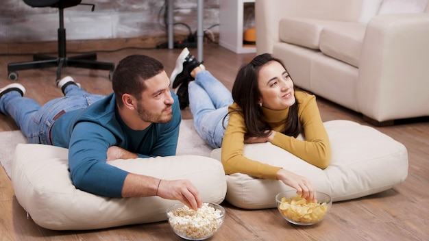 Young woman smiling at his boyfriend while watching tv sitting\
on the floor.