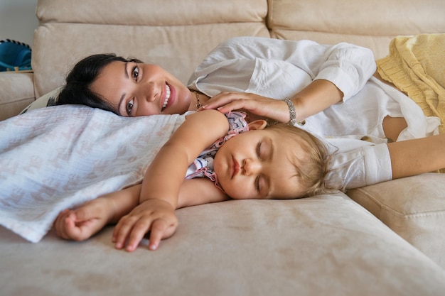 Young woman smiling and looking at camera while lying with cute little daughter sleeping on comfortable sofa in living room at home