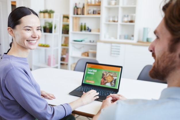 Giovane donna che sorride all'uomo e che fa la consegna del cibo sul laptop mentre sono seduti al tavolo in cucina