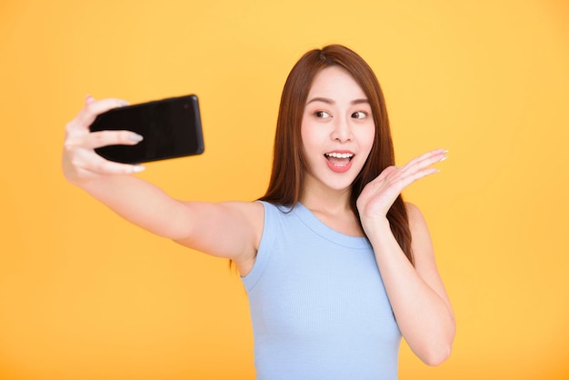 Young woman smiling and taking selfie photo on cellphone isolated over yellow background