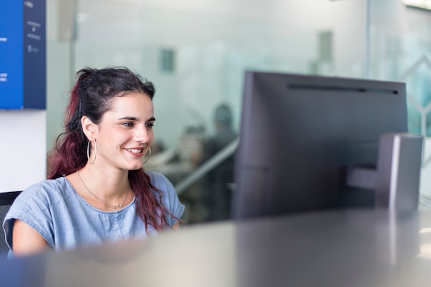 Молодая женщина улыбается, наблюдая сообщения на экране компьютера, в Коворкинг пространстве.