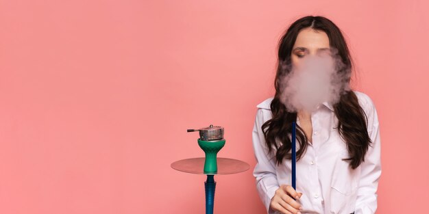 Young woman smokes a hookah, shisha on a pink background. Place for your text.