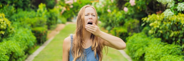Young woman sneezes in the park against the background of a flowering tree. Allergy to pollen concept BANNER long format