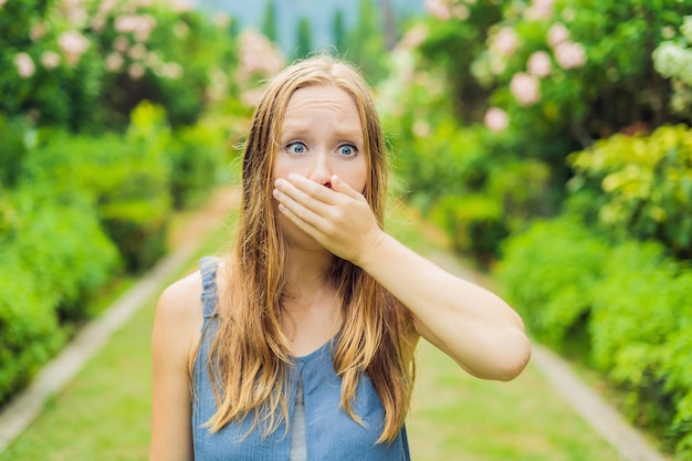 Young woman sneezes in the park against the background of a flowering tree. Allergy to pollen concept