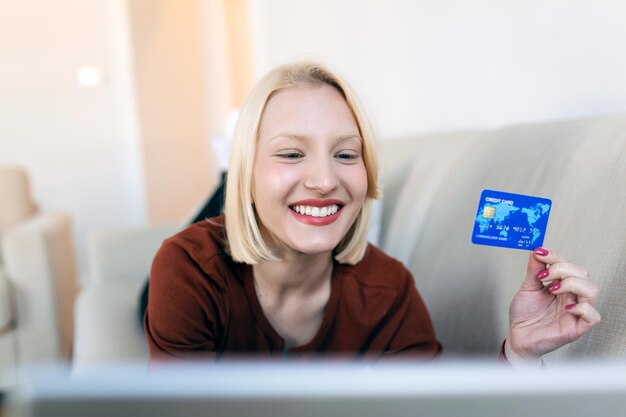 직불 카드로 온라인 쇼핑을 하는 소파에 있는 젊은 여성 집에서 온라인 쇼핑을 위해 노트북 컴퓨터를 사용하는 아름다운 여성
