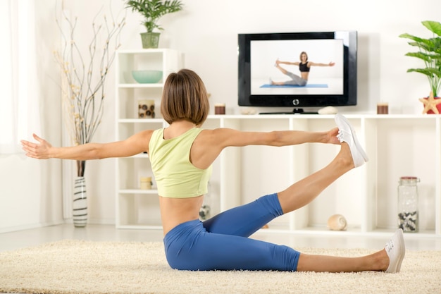 Young woman in sports clothes, doing exercises in the room, in front TV.