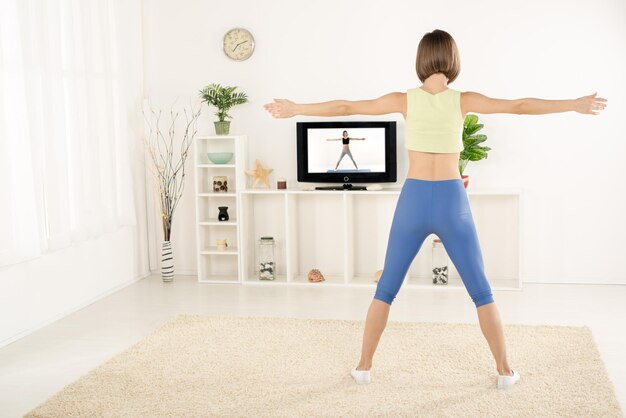 Young woman in sports clothes, photographed from behind, exercise in the room, watching the exercises on TV.