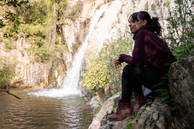 Молодая женщина в спортивной одежде сидит и смотрит на небольшой водопад посреди леса.