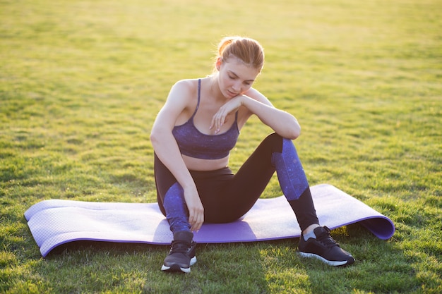 Молодая женщина в спортивной одежде, сидя на тренировочный мат в поле