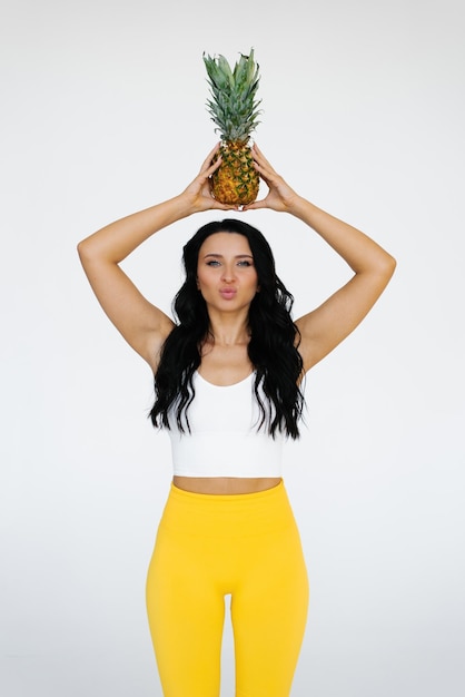 Молодая женщина в спортивной одежде держит ананас над головой и сложила губы, как будто целует концепцию здорового питания