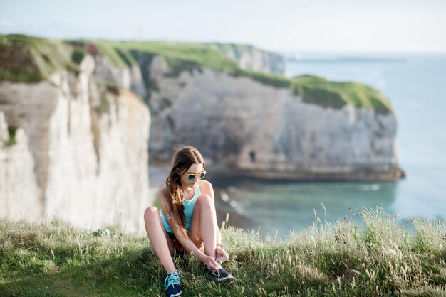 Young woman in sportswear tying shoelaces sitting outdoors on the beautiful rocky coastline background near Etretat town in France