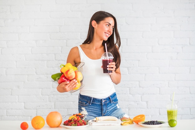 Giovane donna che sta contro la ciotola della tenuta della parete di verdura fresca e frutta e succo