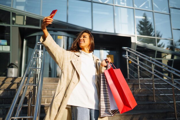 Photo young woman standing along road holding shopping bags using mobile phone discounts sale concept