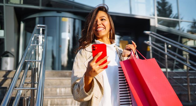 Foto giovane donna in piedi lungo la strada con le borse della spesa usando il telefono cellulare concetto di vendita di sconti