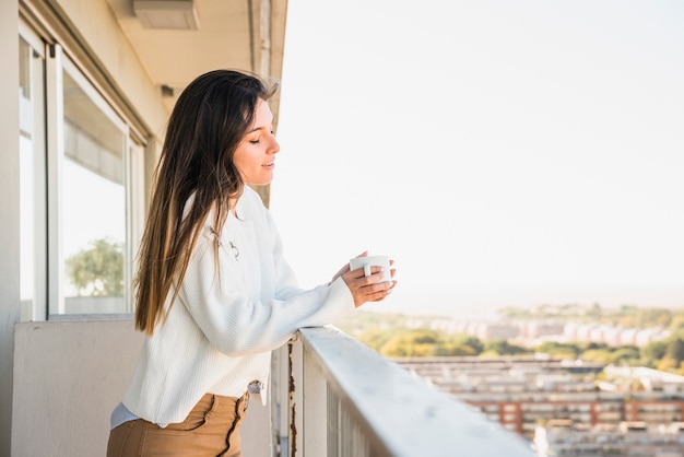 Молодая женщина, стоя на балконе, держа чашку кофе