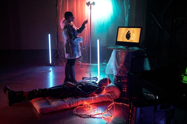 Young woman standing by dropper while young man lying on mattress by computer monitor in dark room