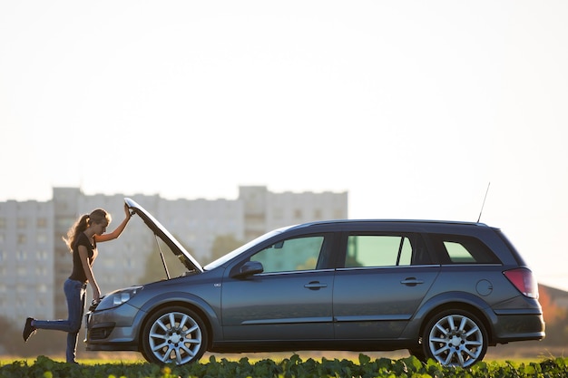 Young woman standing next to car looking under popped hood