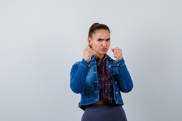 Young woman standing in fight pose in checkered shirt, jean jacket and looking confident , front view.