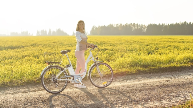 Photo young woman standing next to her electric bike over dusty country road, strong afternoon sun backlight in background shines on yellow flowers field, view from side