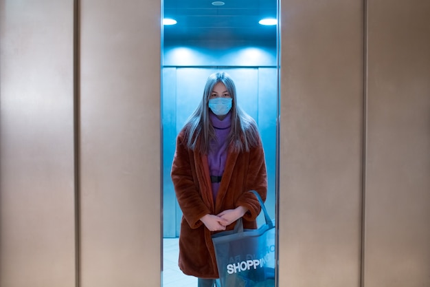 Молодая женщина, стоящая в закрывающемся лифте в медицинской маске для лица, пандемия коронавируса и концепция покупок