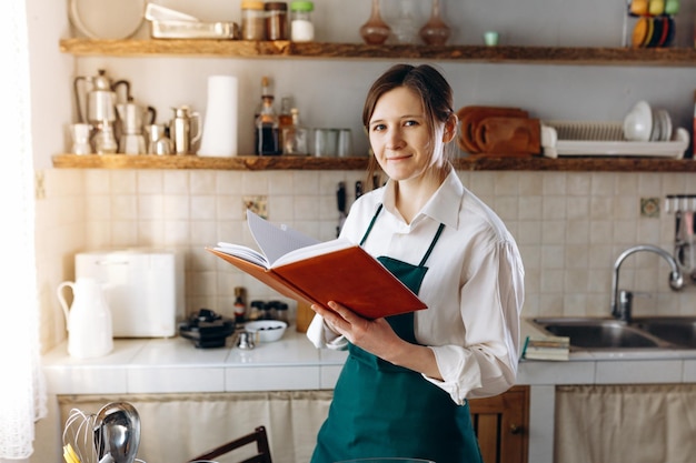 レシピ本を持ってキッチンに立つ若い女性。家庭での料理のコンセプト、ライフスタイル。