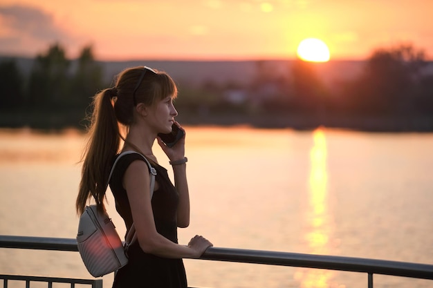 暖かい夜に屋外で彼女の携帯電話で話している夏の公園の湖岸に立っている若い女性コミュニケーションとモバイル接続の概念