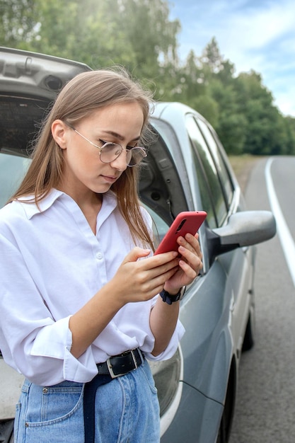 Young woman standing near broken down car with popped up hood having trouble with her vehicle Waiting for help tow truck or technical support A woman calls the service center