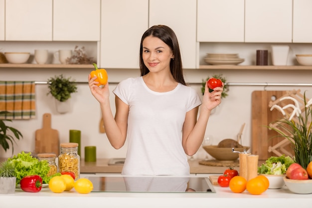 Young woman standing near desk and laptop in the kitchen, smiling, looking at camera, with ingredients.