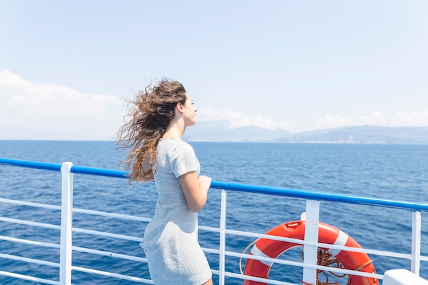 青い海を見て船の手すりの横に立っている若い女性