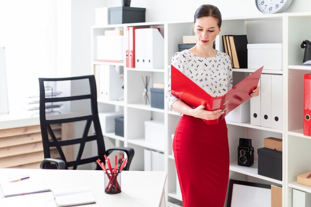 A young woman standing in the office and holding a red folder with documents.