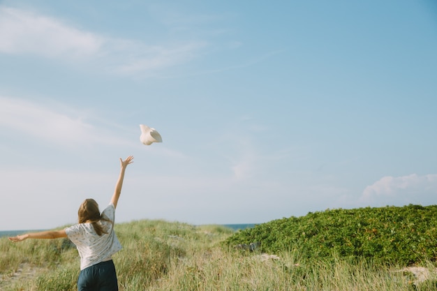 Фото Молодая женщина стоит на травянистом пляже и бросает свою шляпу в небо