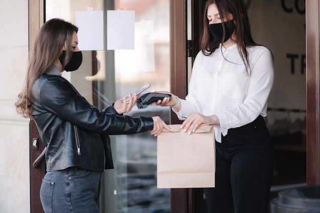 Young woman standing outdoors in mask and paying bill through smartphone using nfc technology in a