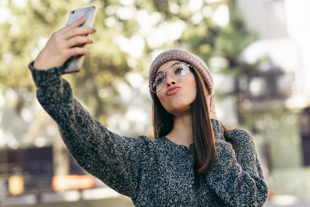 屋外に立ってスマートフォンで自画像を撮る若い女性ニットのセーター帽子の眼鏡を着たブルネットの女性が街の通りで彼女のデバイスで自分撮りを作る