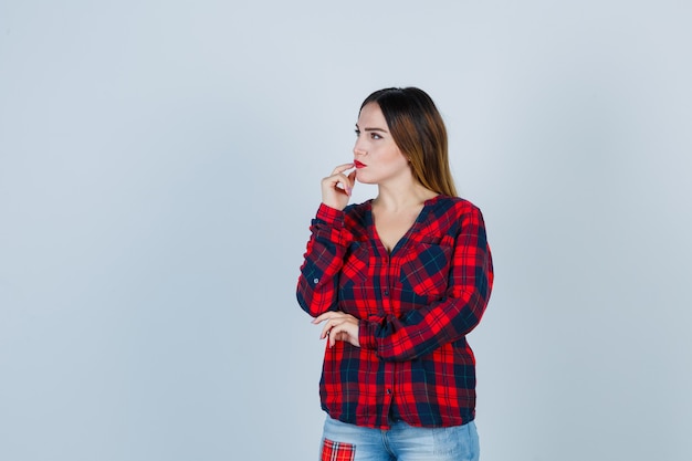 Foto giovane donna in piedi in posa di pensiero, tenendo le dita sulla bocca in camicia a quadri, jeans e guardando pensieroso. vista frontale.