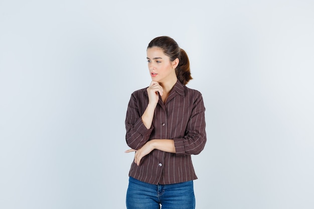 Young woman standing in thinking pose, propping chin on hand in striped shirt, jeans and looking pensive , front view.