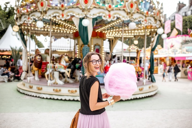 Young woman standing with pink cotton candy outdoors in front of the carrousel at the amusement park