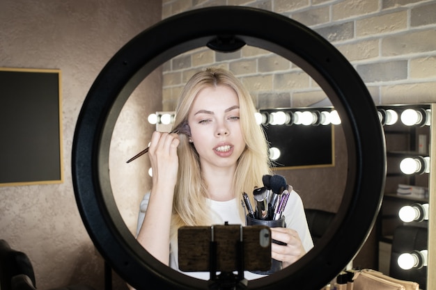 Молодая женщина стоит перед кольцевой лампой и проводит онлайн-курсы по нанесению макияжа.