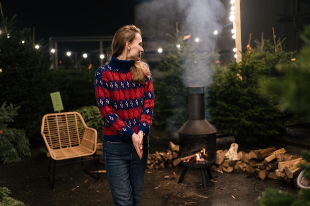 A young woman stands in the middle of Christmas trees and looks at the stove and firewood