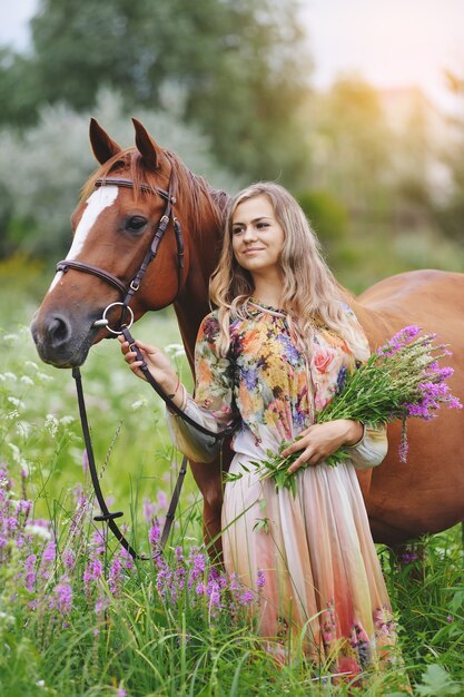 Young woman stands near a horse in a summer dress on a meadow