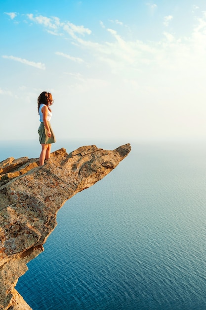 A young woman stands on a picturesque steep cliff above the sea against the sky