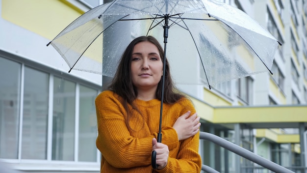 Young woman stands under a transparent umbrella in the rain in cold weather, slow motion