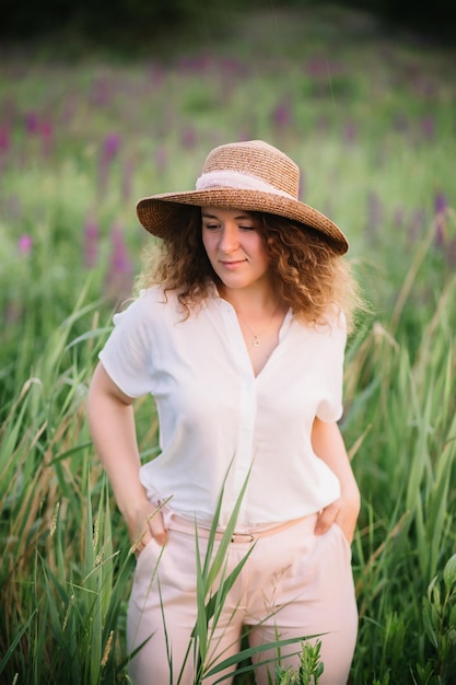 Young woman stands in white shirt in field of purple and pink lupins Beautiful young woman with curly hair and hat outdoors on a meadow lupins blossom Sunset or sunrise bright evening light