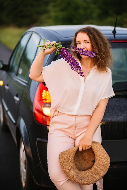 Young woman stands in white shirt near car with purple and pink lupins Beautiful young woman