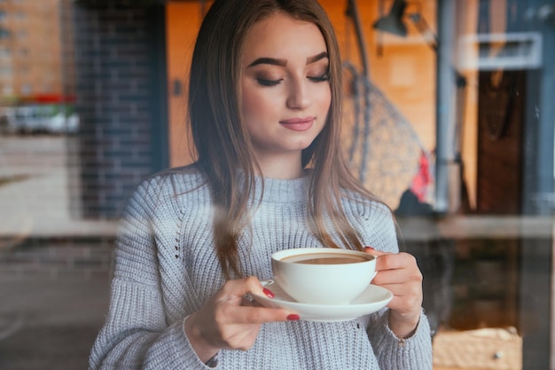 A young woman stands at the window with a cup of coffee City cafe The girl spends her free time in a cozy atmosphere