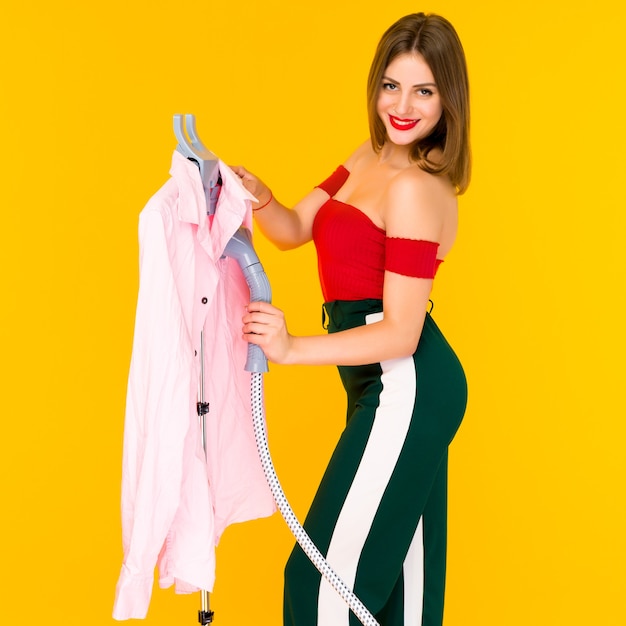 Young woman steaming pink shirt at dry-cleaner's on yellow background