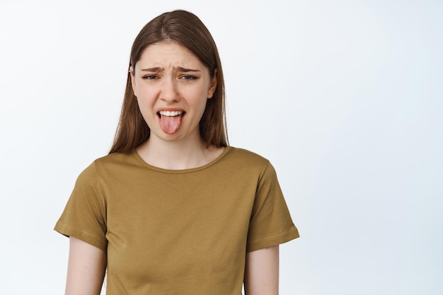 젊은 여성은 싫어서 혀를 내밀고, 나쁜 맛에 역겹고, 끔찍하거나 역겹고, 흰색 배경 위에 서 있습니다.