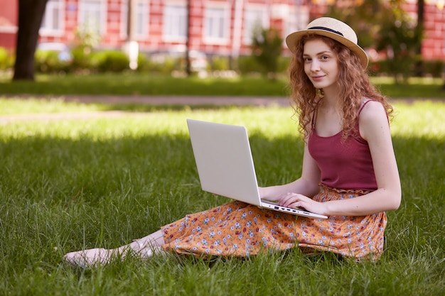 Young woman in straw hat, burgundy t shirt and skirt sitting on green grass and browse in internet with her white laptop, has online classes, attractive female with wavy foxy hair studies inopen air.