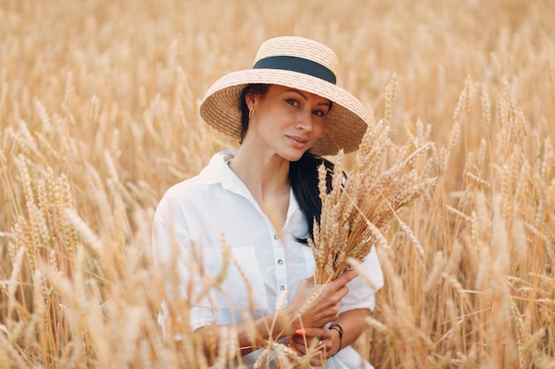 Young Woman in straw hat holding sheaf of wheat ears at agricultural field.